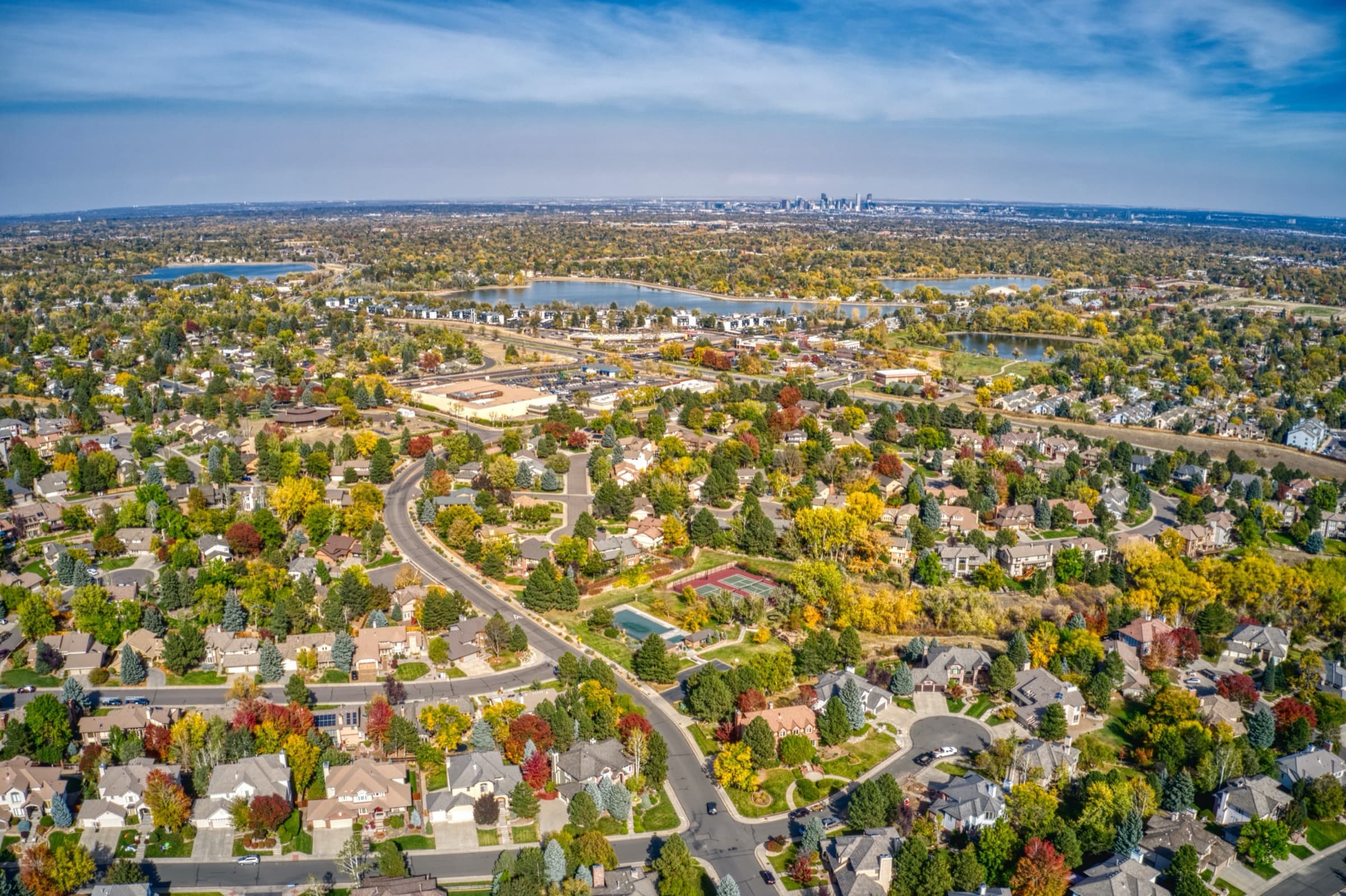 Aerial view of tree-lined neighborhoods and lakes in fall color, with the downtown Denver skyline in the distance